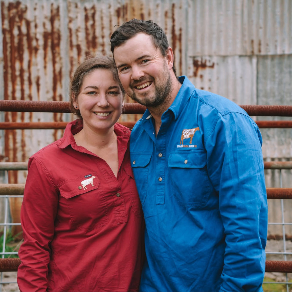 A smiling man and woman standing side by side in front of a rustic corrugated metal wall and rusty fence. The woman wears a red long-sleeve collared shirt with a small white logo on the left chest, and the man wears a bright blue long-sleeve button-up shirt with a small orange and white logo on the left chest.